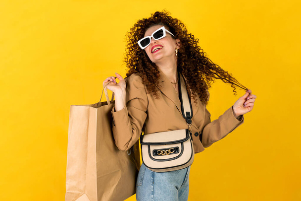 Curly κορίτσι στο μοντέρνο ντύσιμο τζιν και σακάκι ποζάρουν με τσάντα για ψώνια στο κίτρινο φόντο στο στούντιο. Χαρούμενη έκφραση προσώπου γυναίκας. Έννοια αγορών. - Φωτογραφία, εικόνα