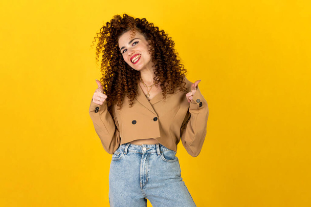 Lockiges Mädchen im trendigen Outfit aus Jeans und Jacke posiert auf gelbem Hintergrund im Studio. Gesichtsausdruck der Frau. Isoliertes Porträt. - Foto, Bild