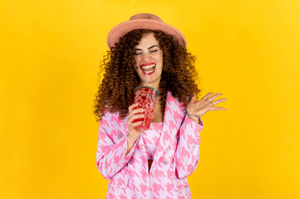 Kırmızı kokteyl içerken pembe giyinen ve eğlenen yaz kıyafetli bir kız. Sarı arkaplanda poz. - Fotoğraf, Görsel