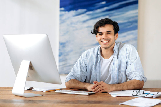 Male successful office employee at workplace. Portrait at work desk of handsome pleasant mixed race hispanic guy, freelancer, manager, looking at camera and smiling friendly - 写真・画像