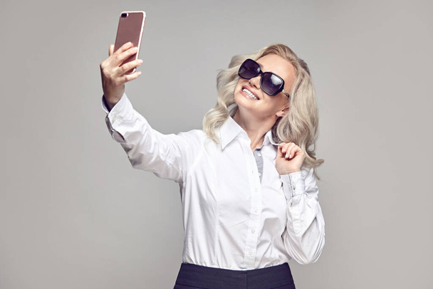 Fröhliche reife Frau mit Sonnenbrille macht ein Selfie, um es in den sozialen Medien auf grau isoliert zu teilen - Foto, Bild