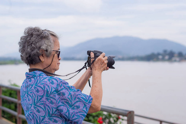 Une femme âgée aux cheveux gris court tenant un appareil photo numérique tout en se tenant au bord de la rivière avec un fond naturel. Café du matin. Concept de personnes âgées et photographie. - Photo, image