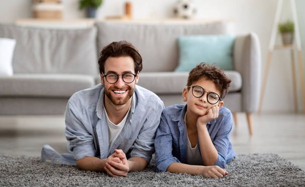 Портрет веселого отца и сына в очках и улыбающихся в камеру, лежащих на ковре на полу - Фото, изображение