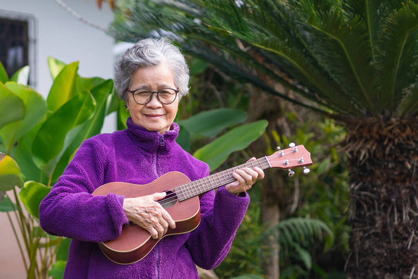 庭に立っている間にウクレレを演奏する高齢女性の肖像画。歌うことでリラックスし、小さなギターの幸せを再生し、引退後の生活をお楽しみください。高齢者と医療の概念. - 写真・画像