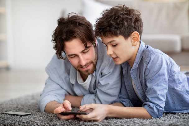 Πατέρας και γιος χρησιμοποιώντας smartphone, βλέποντας φωτογραφίες ή αγόρι βοηθώντας τον μπαμπά με το παιχνίδι, περνώντας χρόνο στο σπίτι - Φωτογραφία, εικόνα