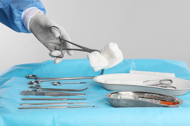 Doctor holding forceps with tissue over surgical instruments on table against light background, closeup - Foto, Imagem