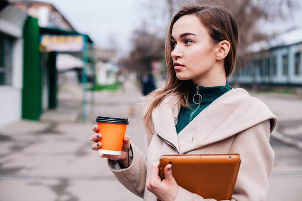 Young, beautiful girl walks down the street with a tablet and coffee in a disposable cup - Zdjęcie, obraz