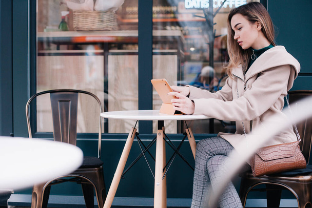 Young, beautiful girl works on a tablet at a street table and drinks coffee from a disposable cup - Foto, Imagem