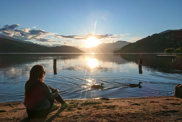 Una mujer sentada a la orilla del lago Millstaetter durante el atardecer. Altos Alpes alrededor. El sol se pone detrás de densas nubes. La superficie tranquila del lago refleja el cielo. Un cisne y un pato balanceándose. - Foto, imagen