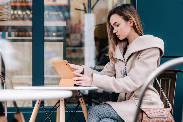 Genç, güzel bir kız sokak masasındaki tablette çalışıyor ve tek kullanımlık bir bardaktan kahve içiyor. - Fotoğraf, Görsel