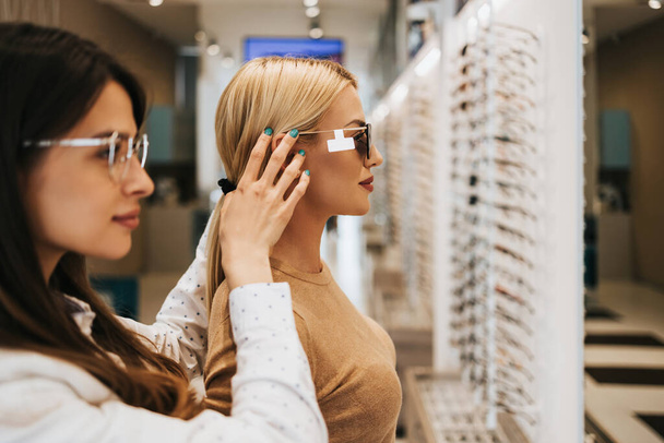 Beautiful and fashionable woman choosing eyeglasses frame in modern optical store. Female seller specialist helps her to make right decision. - Foto, imagen