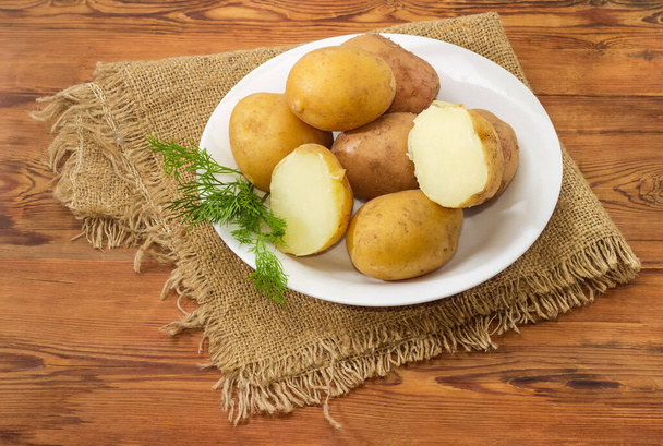 Ziemniaki gotowane w skórkach z gałązką koperkową na białym talerzu na starym rustykalnym stole z workiem - Zdjęcie, obraz