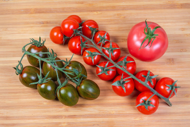 Dos racimos de tomates cherry rojos maduros frescos y tomates cherry kumato, tomate rosado ordinario único en una tabla de cortar de madera, vista superior - Foto, Imagen