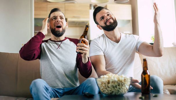Two best friends and fans of football watching some sport match on the TV and drinking beers and eating snacks while cheering for the team on the couch - Fotografie, Obrázek