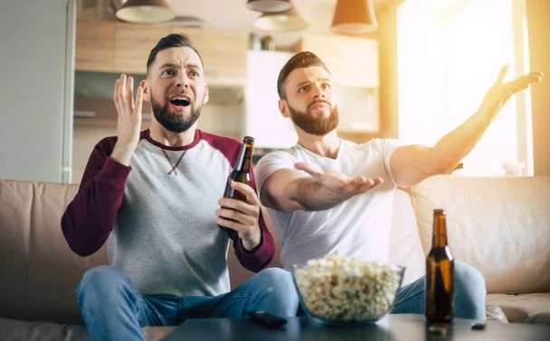 Two best friends and fans of football watching some sport match on the TV and drinking beers and eating snacks while cheering for the team on the couch - Фото, зображення