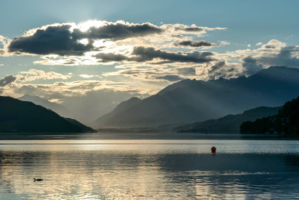 Avusturya 'da Millstaetter Gölü' nde gün batımı. Göl yüksek Alplerle çevrilidir. Gölün sakin yüzeyi güneş ışınlarını yansıtıyor. Güneş dağların arkasında batıyor. Biraz bulutlu. Doğal güzellik. - Fotoğraf, Görsel