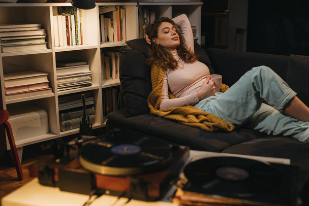 nainen rentoutuu sohvalla kotonaan ja kuuntelee musiikkia levysoitin - Valokuva, kuva