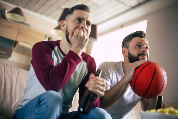 Zwei aufgeregte, lustige junge Basketballfans, die sich ein Fernsehspiel anschauen und auf der Couch ausruhen - Foto, Bild