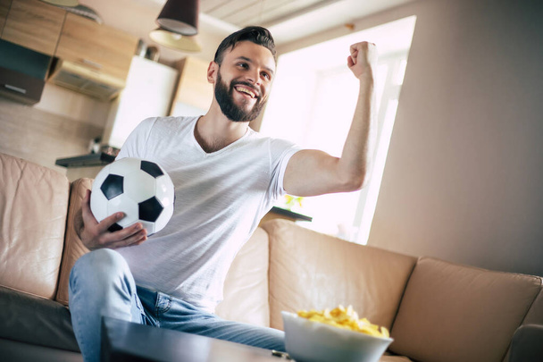 Handsome young excited modern bearded man is relaxing on the couch while watching football match on TV - Foto, afbeelding