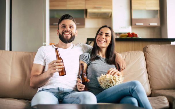 Beautiful smiling modern couple in love is watching some film or movie on TV while they relaxing on the couch at own apartment - Foto, Imagem