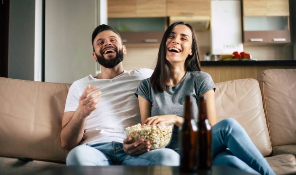 Beau sourire couple moderne amoureux est de regarder un film ou un film à la télévision pendant qu'ils se détendre sur le canapé dans son propre appartement - Photo, image