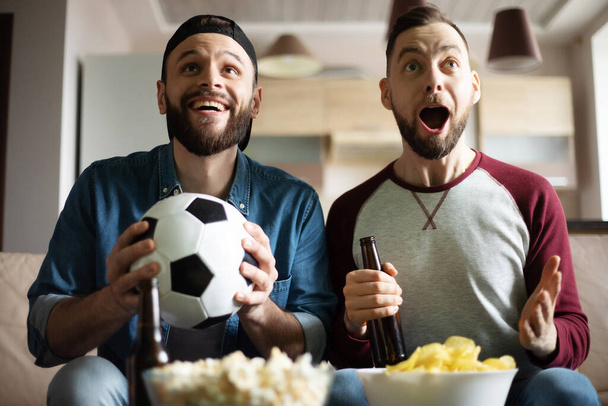 Funny excited hipster bearded friends with snacks and beers watching football on tv while sitting on the couch at home. Fans shouting due to winning of them team - Foto, imagen