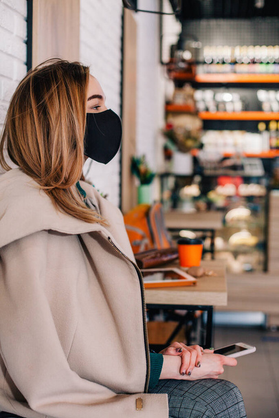 Young girl in protective mask sits in a coffee shop waiting for a drink - Foto, imagen