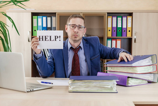 Boss businessman at his desk in the office with a bunch of folders with papers. Holds a HELP sign in his hands. Strenuous office work. Preparing of report - Photo, Image