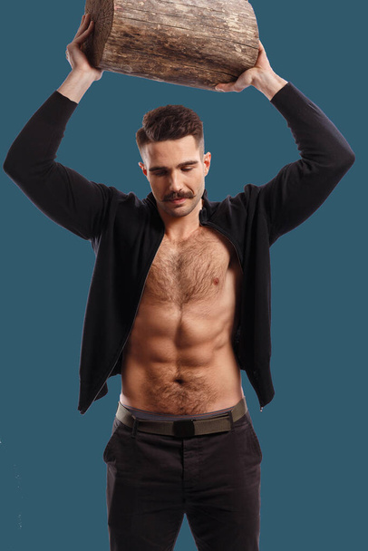 Frontal portrait of a fit, athletic, sexy man with moustache, with naked torso, showing six pack abs, rise up a tree trunk above the head, looking down, over green background. - Photo, Image