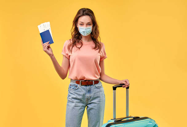 Hübsches Mädchen in lässiger Kleidung mit Koffer, Pass, Tickets und medizinischer Schutzmaske auf gelbem Hintergrund. Tourismus, Reisen, Freizeitkonzept. - Foto, Bild