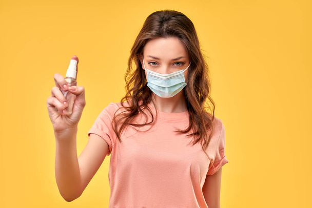 Belle fille en vêtements décontractés et un masque de protection médicale tenant un spray antiseptique isolé sur un fond jaune. - Photo, image