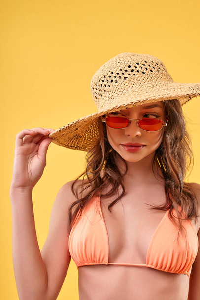 Cute sexy woman in swimsuit, straw hat and sunglasses isolated on yellow background. Travel, vacation, tourism concept. - Photo, Image