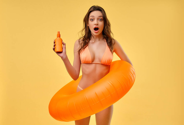 Sexy woman in a swimsuit with an inflatable ring holding a sunscreen isolated on a yellow background. Summer time. Leisure, travel, tourism. - Фото, изображение