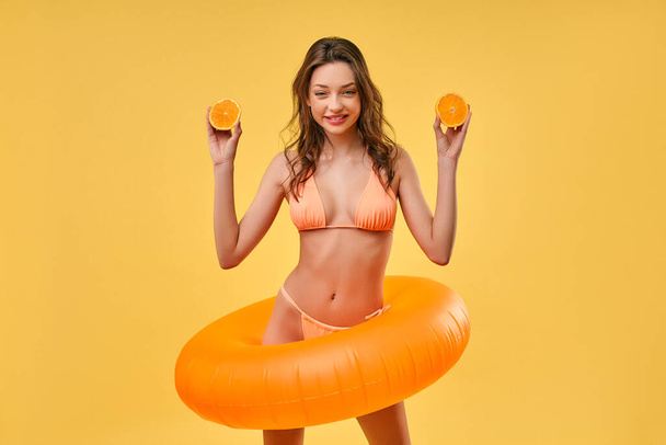 Roztomilá žena v plavkách a s nafukovacím kroužkem, držící půlky pomeranče izolované na žlutém pozadí. Léto. Odpočinek, dovolená, cestování. - Fotografie, Obrázek