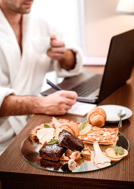 Room service breakfast. Man with beard and in white coat works during breakfast at the hotel. Freelancer. Distant work. Breakfast in the hotel. Close up. Space - Foto, afbeelding