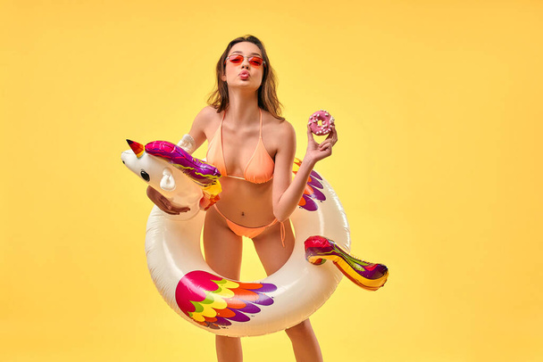 Leuke vrouw in badpak met eenhoorn opblaasbare veelkleurige cirkel en roze donut geïsoleerd op gele achtergrond. Zomertijd. Rust, vakantie, reizen. - Foto, afbeelding