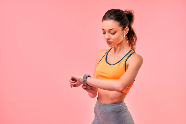 Cute sporty girl in sportswear ticks the time on the clock and listens to music on headphones isolated on a pink background. Sports, yoga, active lifestyle, exercise for health. - Fotoğraf, Görsel