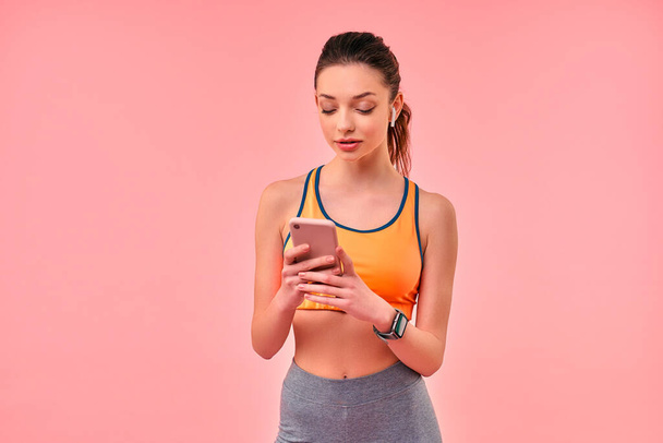 Cute sporty girl in sportswear with phone and wireless headphones isolated on pink background. Sports, yoga, active lifestyle, exercise for health. - Foto, Imagem