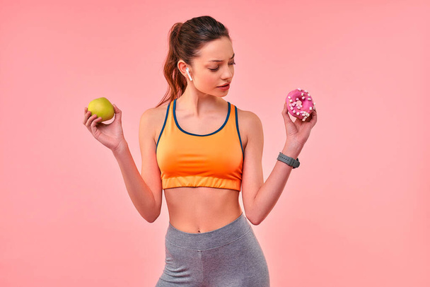 Closeup slim sexy fitness girl hold apple and donut cake in hands. Young beautiful sports woman deciding to choose between healthy fruit or sweet snack. Healthy lifestyle, fitness dieting, weightloss - Photo, Image