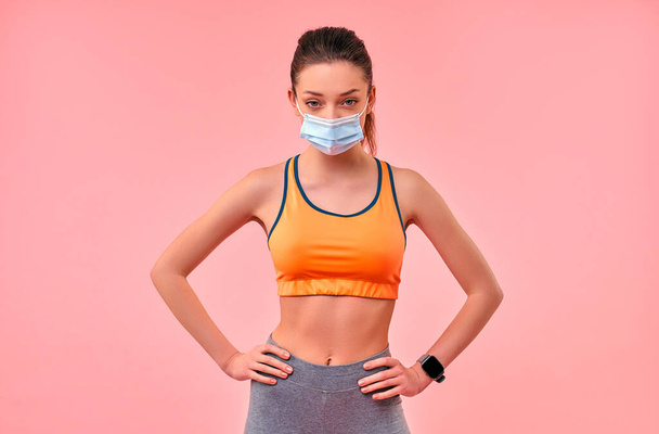 Cute sporty girl in sportswear in a protective medical mask isolated on a pink background. Sports, yoga, active lifestyle in quarantine conditions. - Фото, изображение