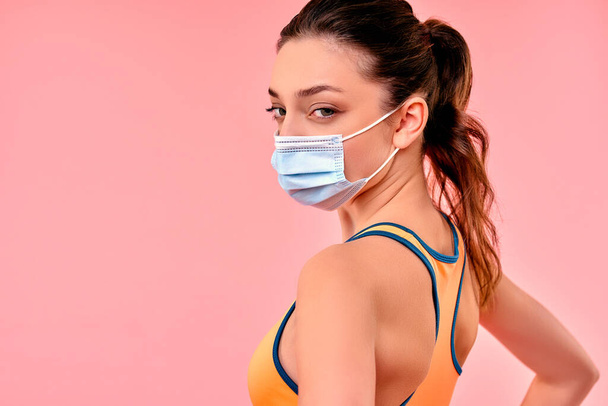 Cute sporty girl in sportswear in a protective medical mask isolated on a pink background. Sports, yoga, active lifestyle in quarantine conditions. - Фото, зображення