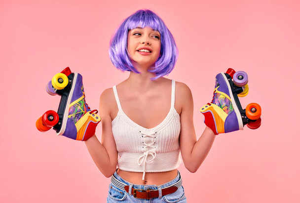 Beautiful young sexy girl in a purple wig is having fun and holding multi-colored roller skates isolated on a pink background. - Photo, image