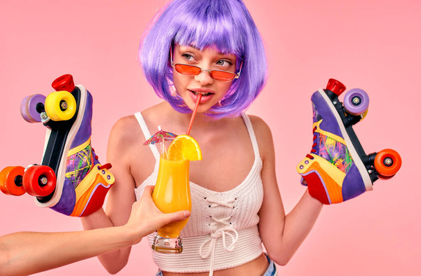 Beautiful young sexy girl in a purple wig is having fun and holding multi-colored roller skates isolated on a pink background. - Фото, зображення