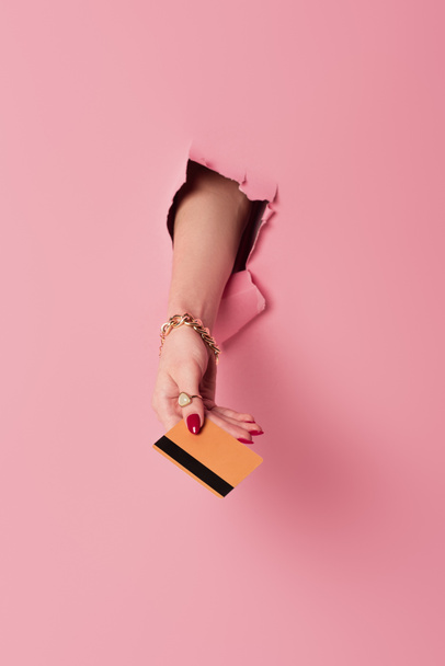 Vue recadrée de la femme avec bracelet et bague tenant la carte de crédit dans le trou sur fond rose - Photo, image