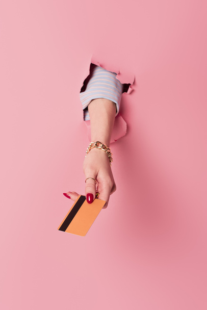 Gewassen weergave van vrouw met rode manicure houden credit card in gat van roze achtergrond  - Foto, afbeelding