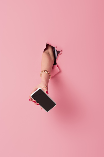 Vista recortada de teléfono inteligente con pantalla en blanco en la mano de la mujer cerca de fondo rosa con agujero  - Foto, Imagen