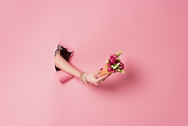 Vue recadrée d'une jeune femme tenant des roses dans un cône de gaufre près d'un fond rose avec trou  - Photo, image
