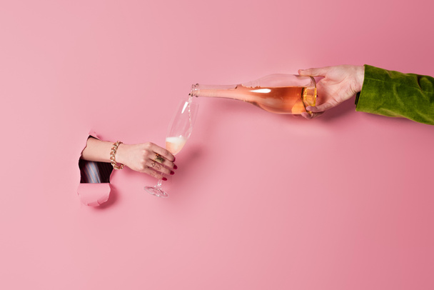 穴のあるガラスとピンクの背景を持つ女性の近くにシャンパンを注ぐ男の作物ビュー  - 写真・画像
