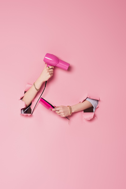 Vista recortada de la mujer sosteniendo peine y secador de pelo cerca de fondo rosa con agujeros  - Foto, Imagen
