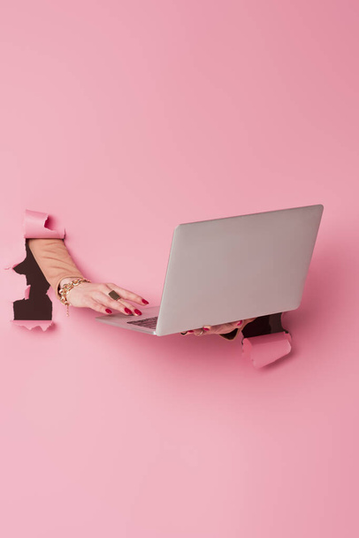 Vista parcial de la mujer con accesorios usando el ordenador portátil cerca de fondo rosa con agujeros  - Foto, Imagen